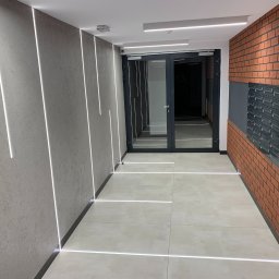 Klaudia Grzesik L-KG Instalacje Elektryczne - Nowoczesny korytarz z oświetleniem LED wzdłuż ścian i podłogi, ceglana ściana z czarnymi skrzynkami pocztowymi, szare drzwi wejściowe.