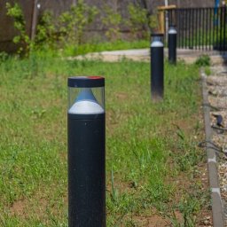 Klaudia Grzesik L-KG Instalacje Elektryczne - Oświetlenie ogrodowe: czarne lampy-słupki z przezroczystymi kloszami, rozmieszczone na trawniku wzdłuż ścieżki z kamyków. Widoczne kable i zieleń w tle.