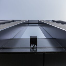 Klaudia Grzesik L-KG Instalacje Elektryczne - Czarny czujnik na elewacji budynku w Makocicach, widok z dołu. Minimalistyczny design, nowoczesna architektura i błękitne niebo w tle. Detal instalacji na fasadzie.