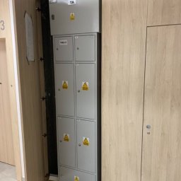 Klaudia Grzesik L-KG Instalacje Elektryczne - Szara szafa elektryczna TA0/2 z ostrzeżeniami, umieszczona w drewnianej wnęce w korytarzu, widoczna z otwartymi drzwiami szafy.