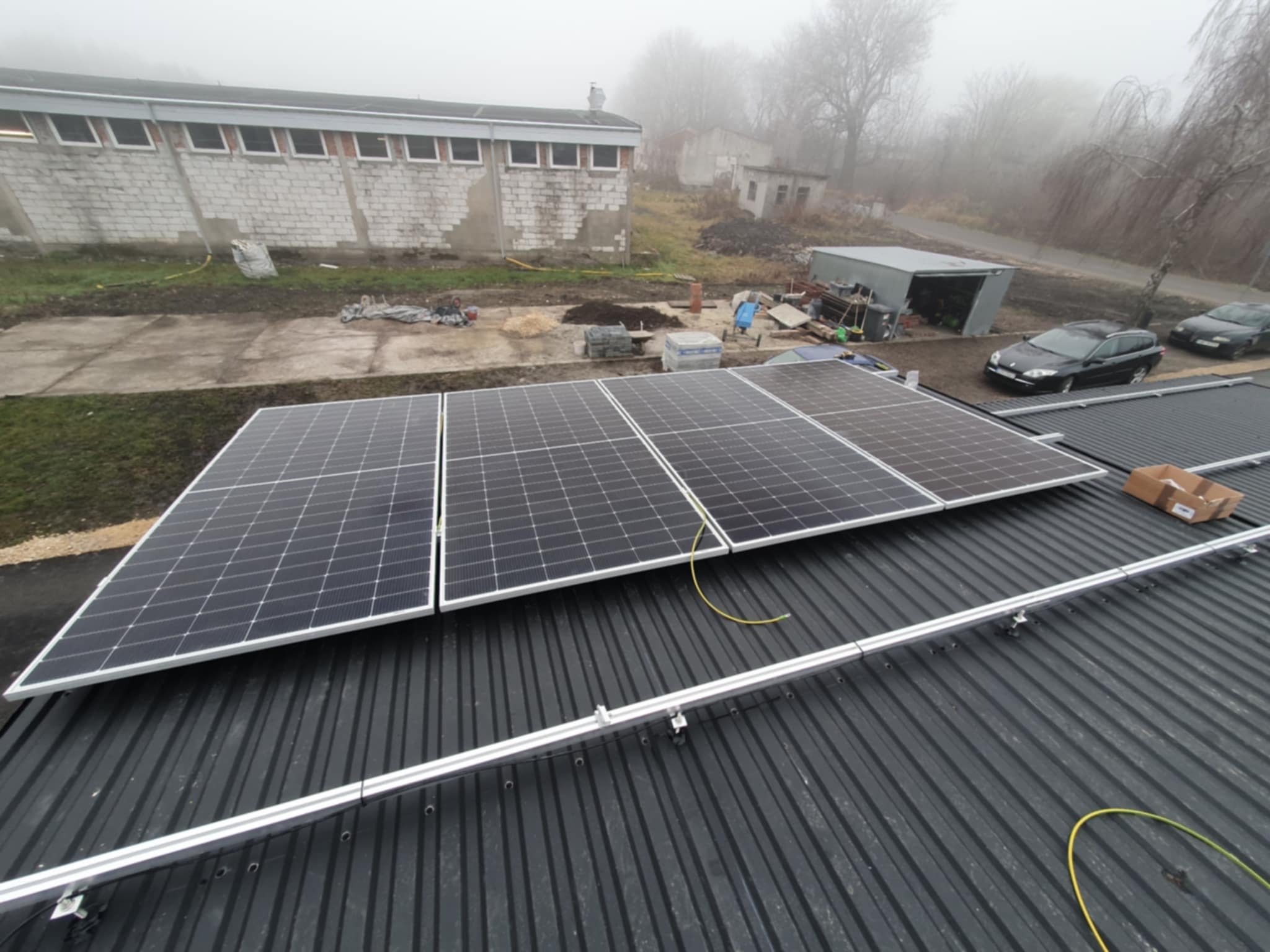 Cztery panele słoneczne zamontowane na dachu z blachy trapezowej, widok z góry na tle zamglonego krajobrazu z budynkami i samochodami.