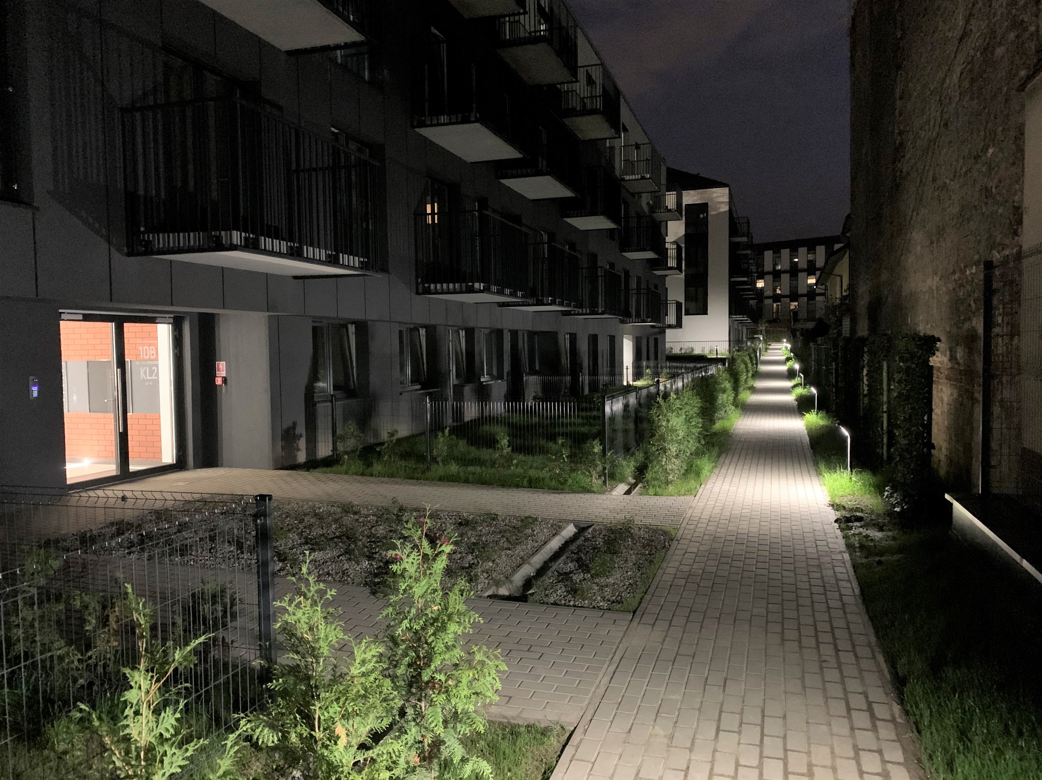 Oświetlona ścieżka prowadząca wzdłuż nowoczesnego budynku mieszkalnego nocą. Lampy oświetlają chodnik, tworząc przyjemną atmosferę.