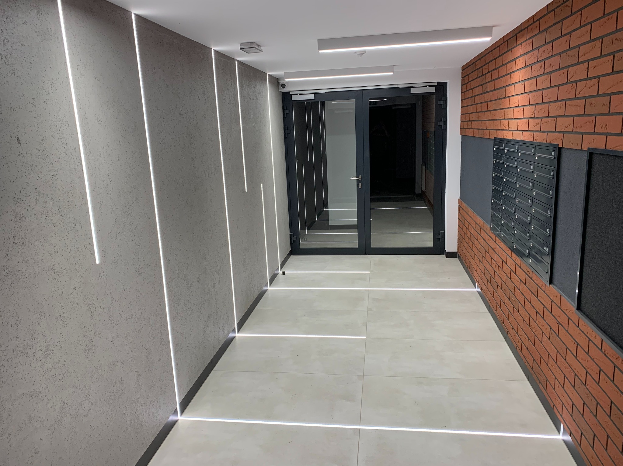 Nowoczesny korytarz z oświetleniem LED wzdłuż ścian i podłogi, ceglana ściana z czarnymi skrzynkami pocztowymi, szare drzwi wejściowe.