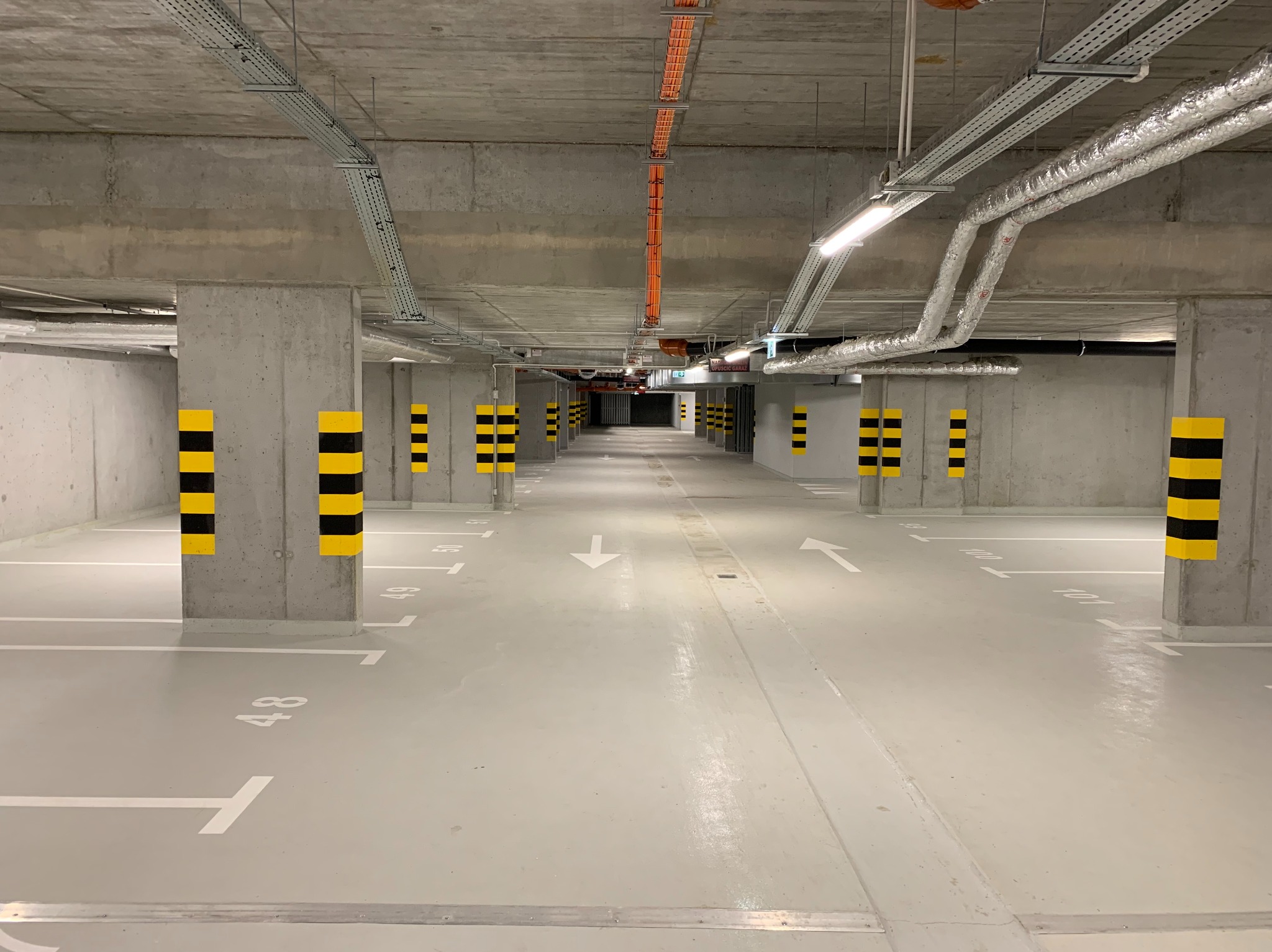 Wnętrze garażu podziemnego z widocznymi instalacjami pod sufitem, oznaczonymi miejscami parkingowymi i słupami z pasami ostrzegawczymi. Perspektywa w głąb garażu.