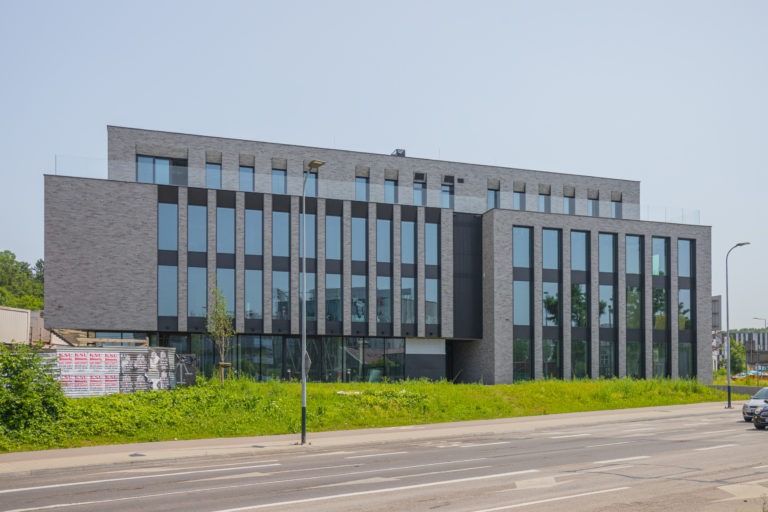 Nowoczesny budynek biurowy z szarej cegły z dużymi oknami, widoczny z poziomu ulicy, z zielenią w tle i słupem oświetleniowym na pierwszym planie.