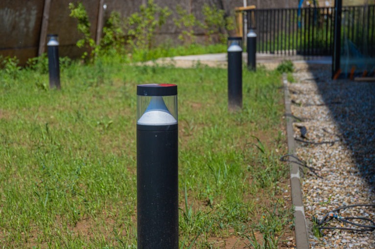 Oświetlenie ogrodowe: czarne lampy-słupki z przezroczystymi kloszami, rozmieszczone na trawniku wzdłuż ścieżki z kamyków. Widoczne kable i zieleń w tle.