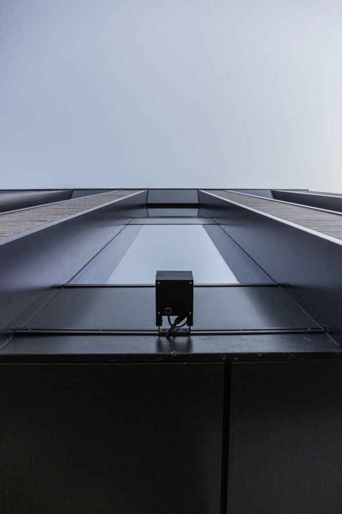 Czarny czujnik na elewacji budynku w Makocicach, widok z dołu. Minimalistyczny design, nowoczesna architektura i błękitne niebo w tle. Detal instalacji na fasadzie.