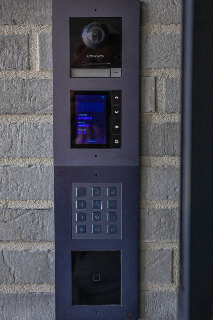 Panel domofonowy Hikvision na ceglanej ścianie. Widoczny ekran, klawiatura numeryczna i czytnik kart. Nowoczesny design, szara obudowa.