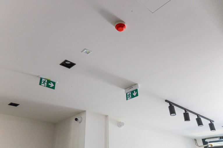 Biały sufit z zamontowanymi elementami: czujnik dymu, oświetlenie punktowe, lampa szynowa, kamera monitoringu i znaki ewakuacyjne z zielonym piktogramem.