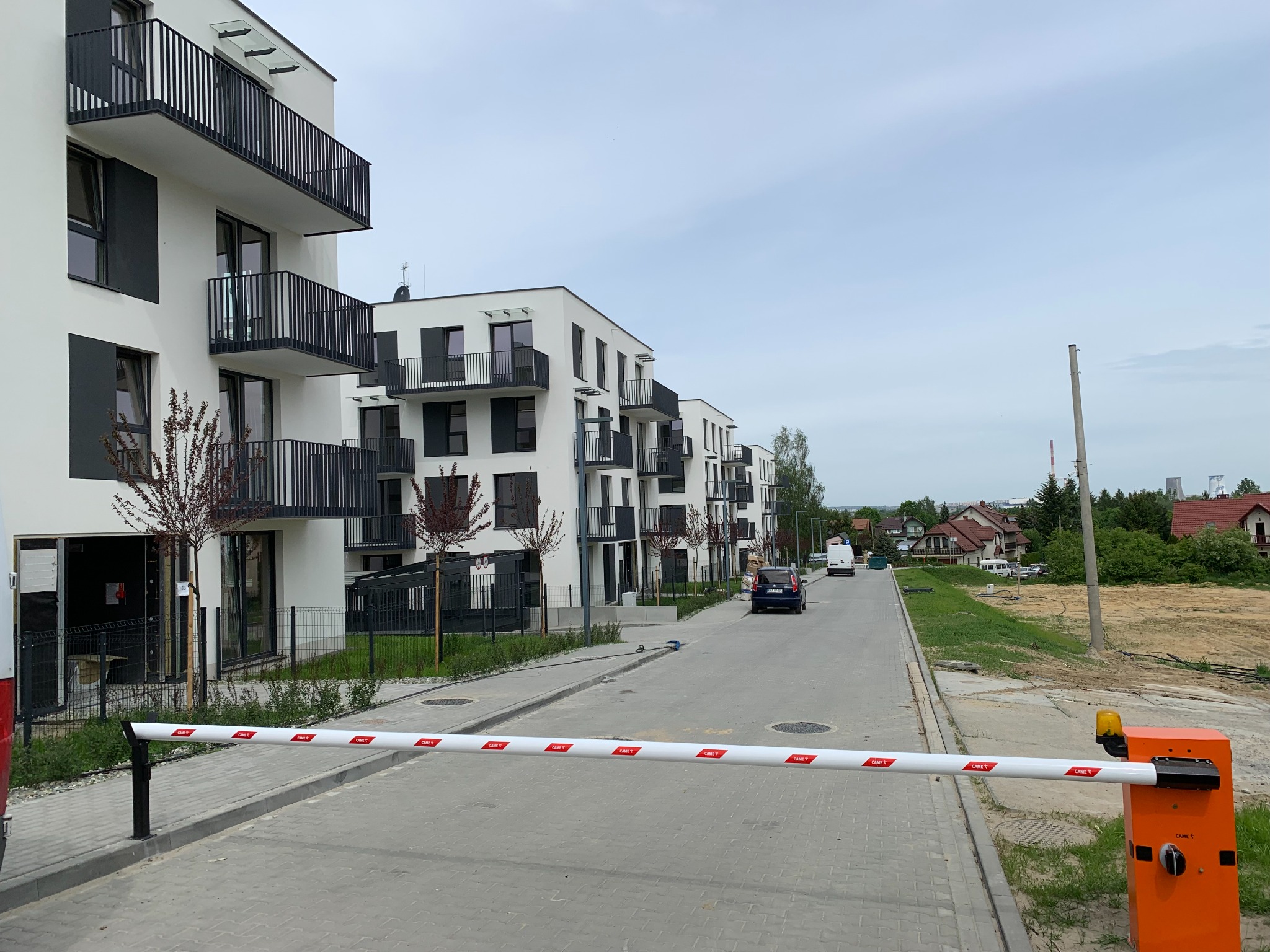 Nowoczesne, białe budynki wielorodzinne z balkonami, widoczne zza szlabanu wjazdowego na osiedle. Ulica z kostki brukowej, w tle zieleń i zabudowa jednorodzinna.