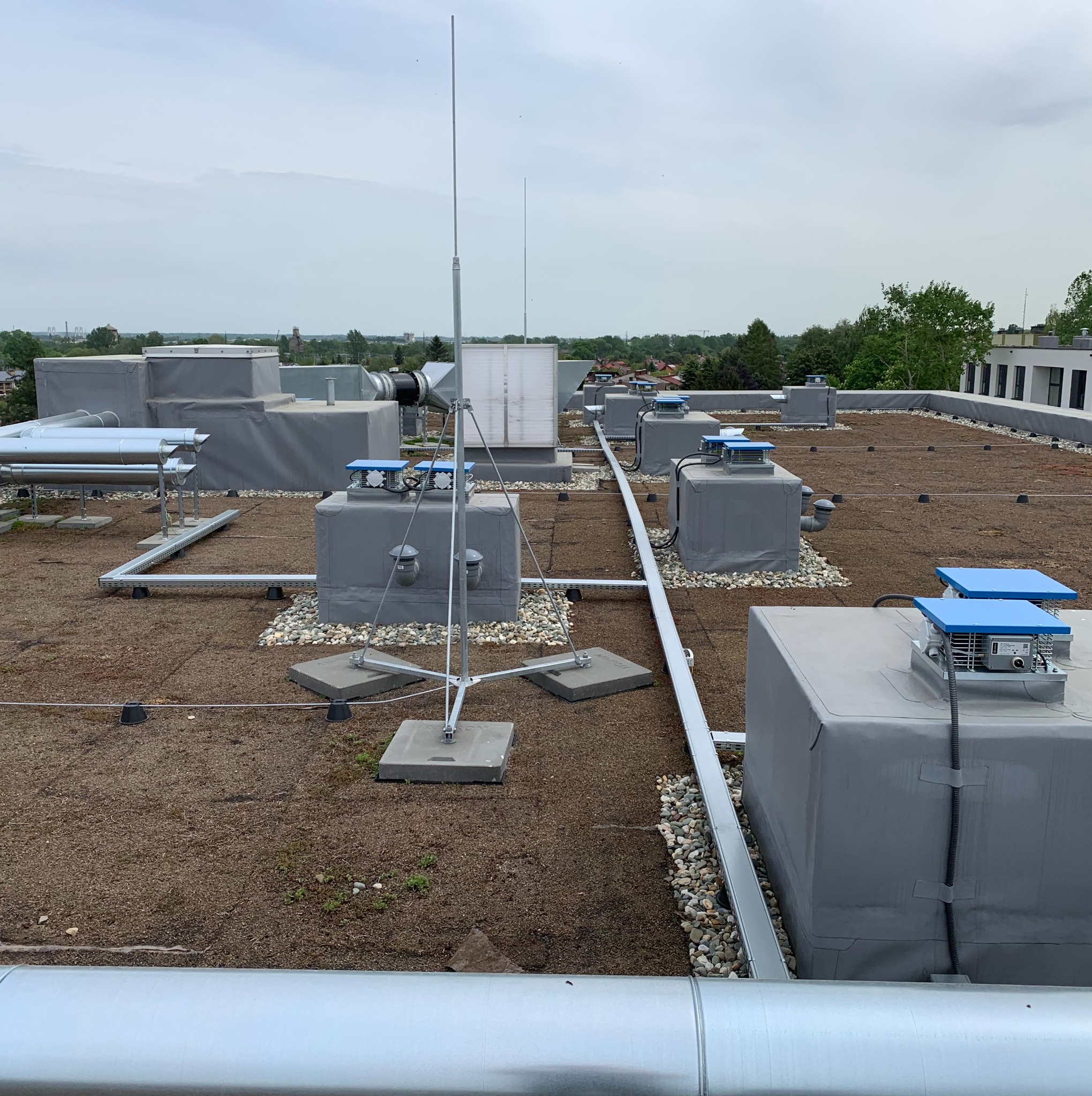 Widok z góry na płaski dach z instalacją odgromową, wentylacją i rurami. Elementy szare, niebieskie i srebrne na tle zielonego dachu. W tle zabudowa miejska i drzewa.