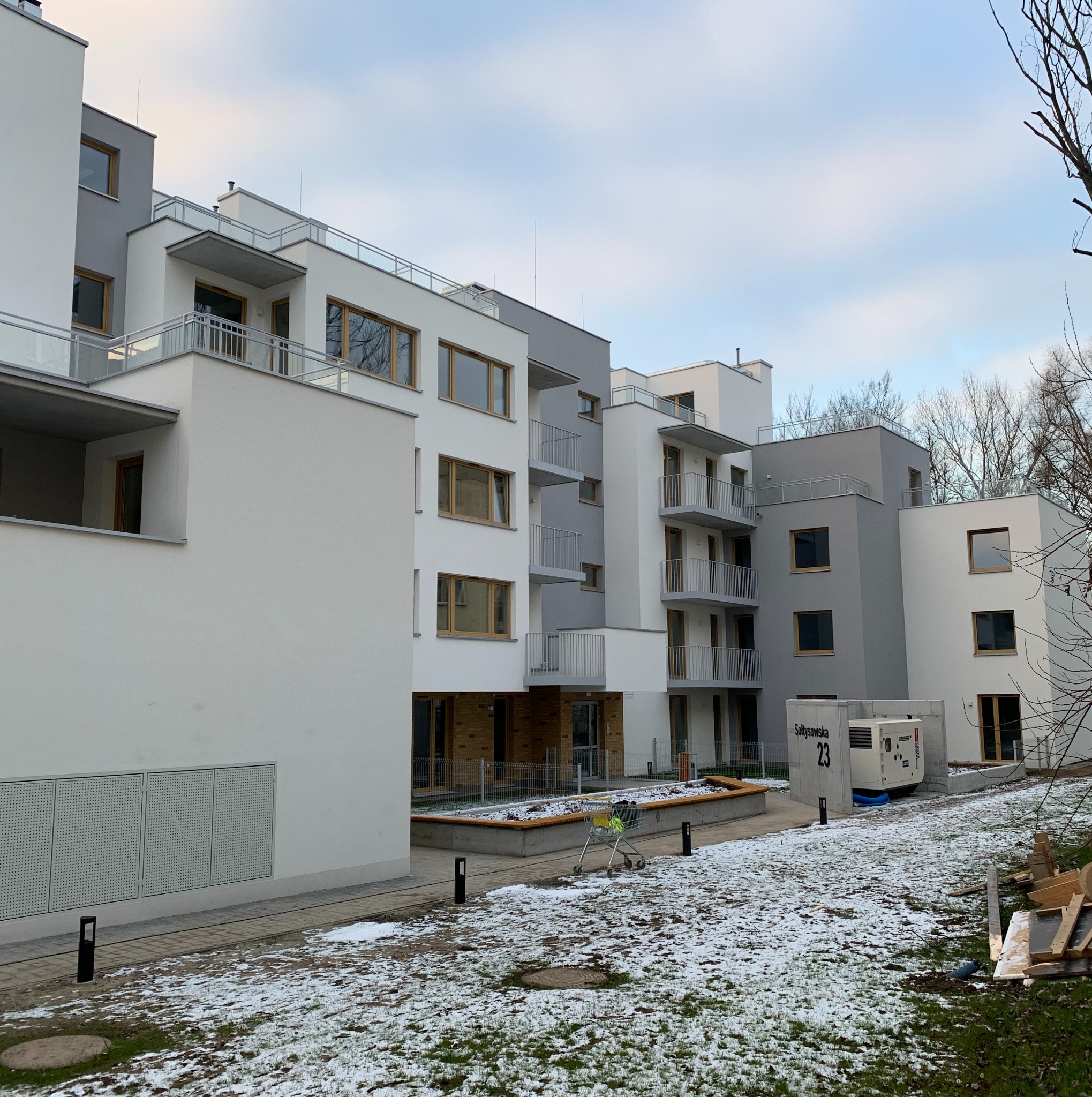 Nowoczesny budynek wielorodzinny z balkonami, w zimowej scenerii. Widoczny agregat prądotwórczy z numerem budynku. Minimalistyczna architektura, biało-szara kolorystyka.