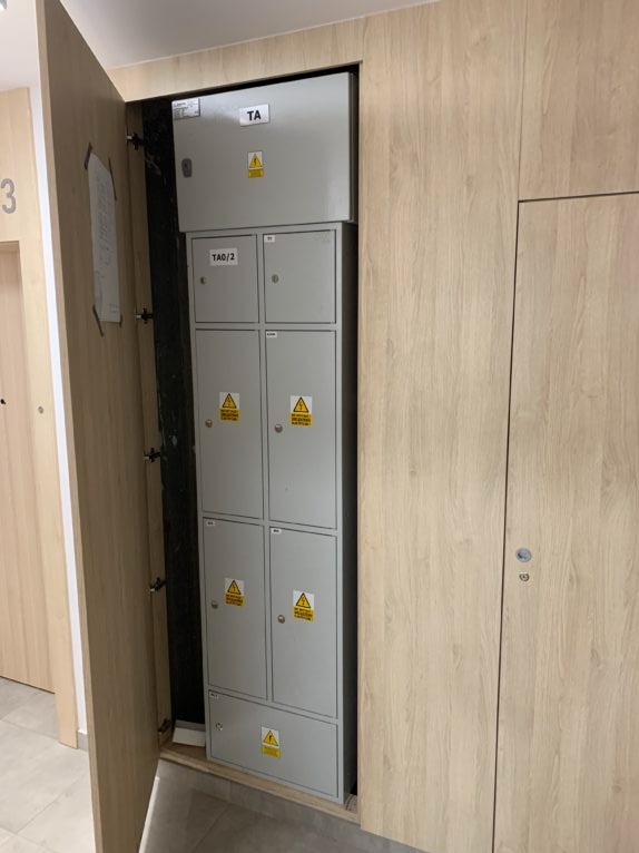 Szara szafa elektryczna TA0/2 z ostrzeżeniami, umieszczona w drewnianej wnęce w korytarzu, widoczna z otwartymi drzwiami szafy.