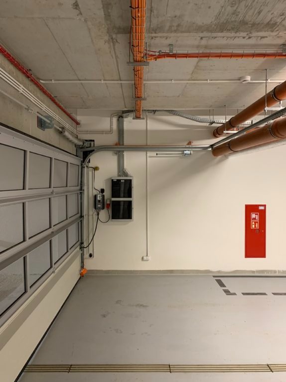 Instalacje w garażu podziemnym: kable w korytach, rury wentylacyjne, skrzynka elektryczna, hydrant. Widok na sufit i ścianę z bramą garażową.