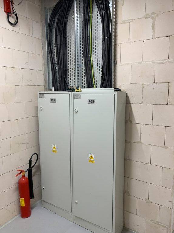 Dwie szafy elektryczne RGA i RGB z przewodami w korytkach, zamontowane w pomieszczeniu z surowych bloczków. Czerwona gaśnica obok. Widoczne znaki ostrzegawcze.