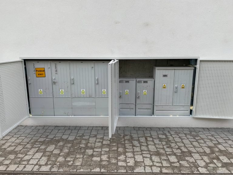 Szara szafka elektryczna z otwartymi drzwiami na zewnątrz budynku, widoczne oznaczenia ostrzegawcze i napisy informacyjne. Widok z bliska na instalację elektryczną.