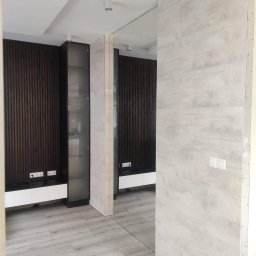 Ivan - Wnętrze mieszkania po remoncie: drewniana boazeria na ścianie, białe meble, szklana witryna, podłoga imitująca drewno, ściana wykończona płytami gipsowo-kartonowymi przed malowaniem, widoczne...