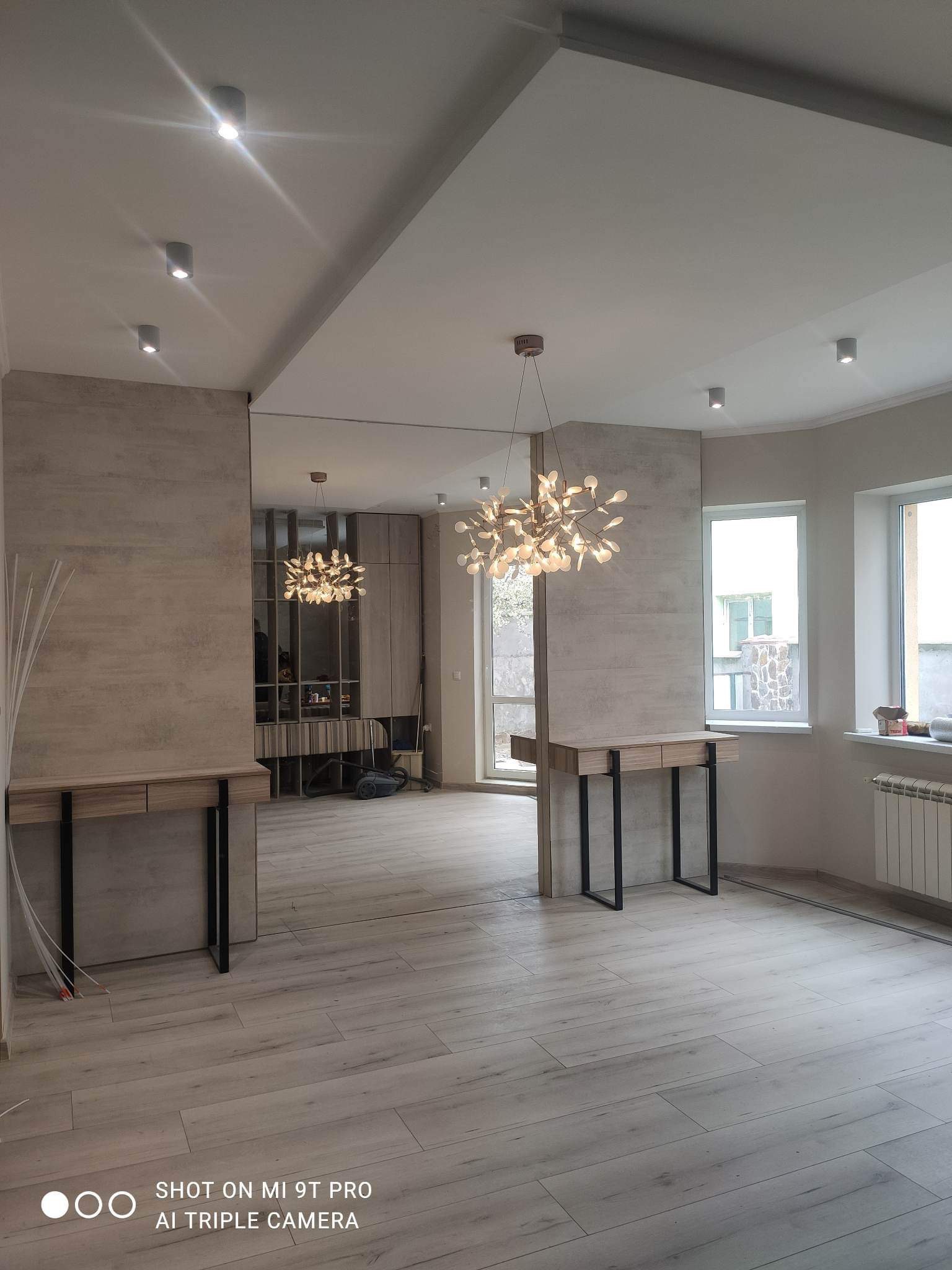 Nowoczesne wnętrze po remoncie z drewnopodobną podłogą, dekoracyjną lampą sufitową, konsolami i lustrem na ścianie, widoczne grzejniki i okna.