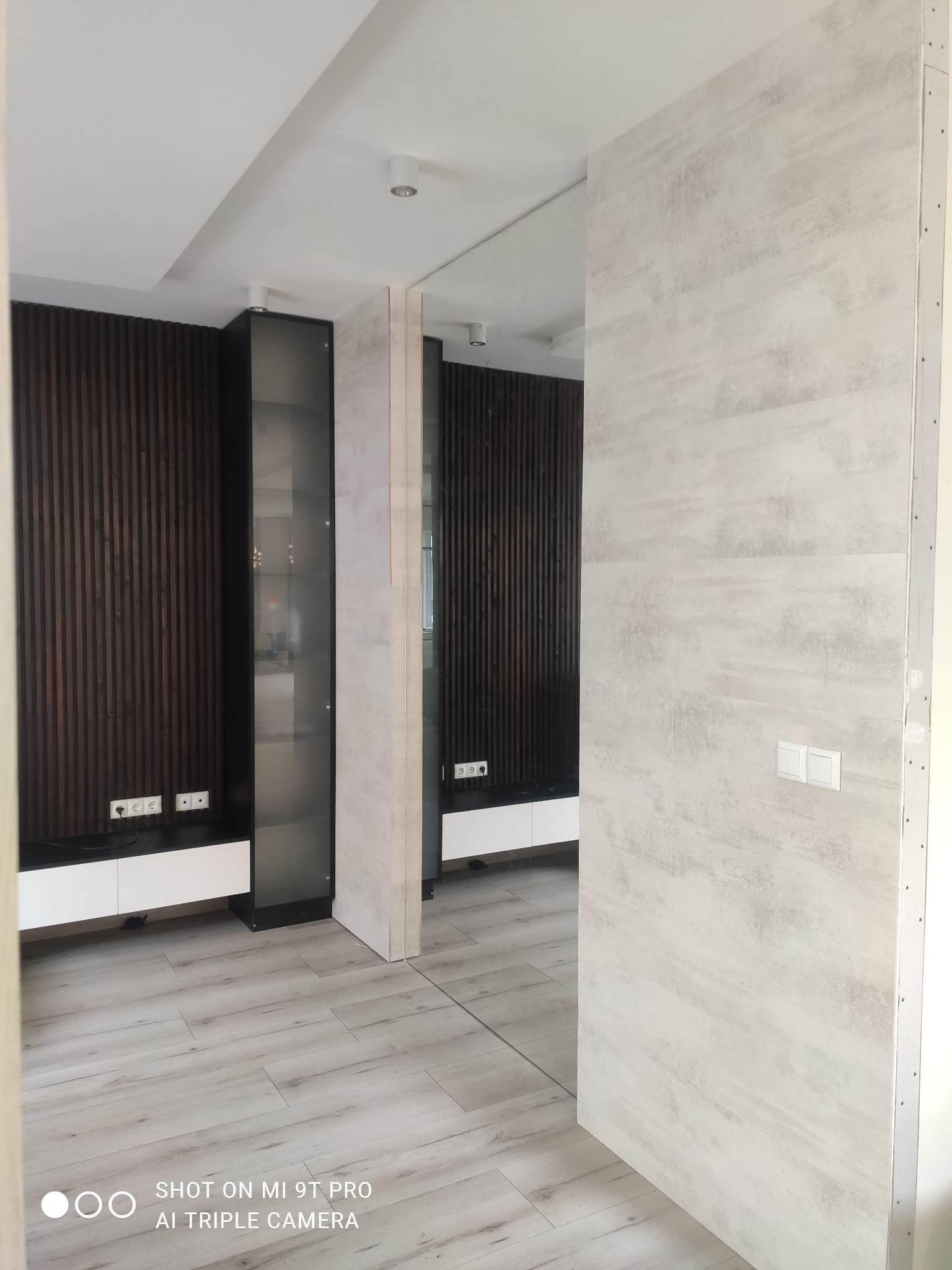 Wnętrze mieszkania po remoncie: drewniana boazeria na ścianie, białe meble, szklana witryna, podłoga imitująca drewno, ściana wykończona płytami gipsowo-kartonowymi przed malowaniem, widoczne...