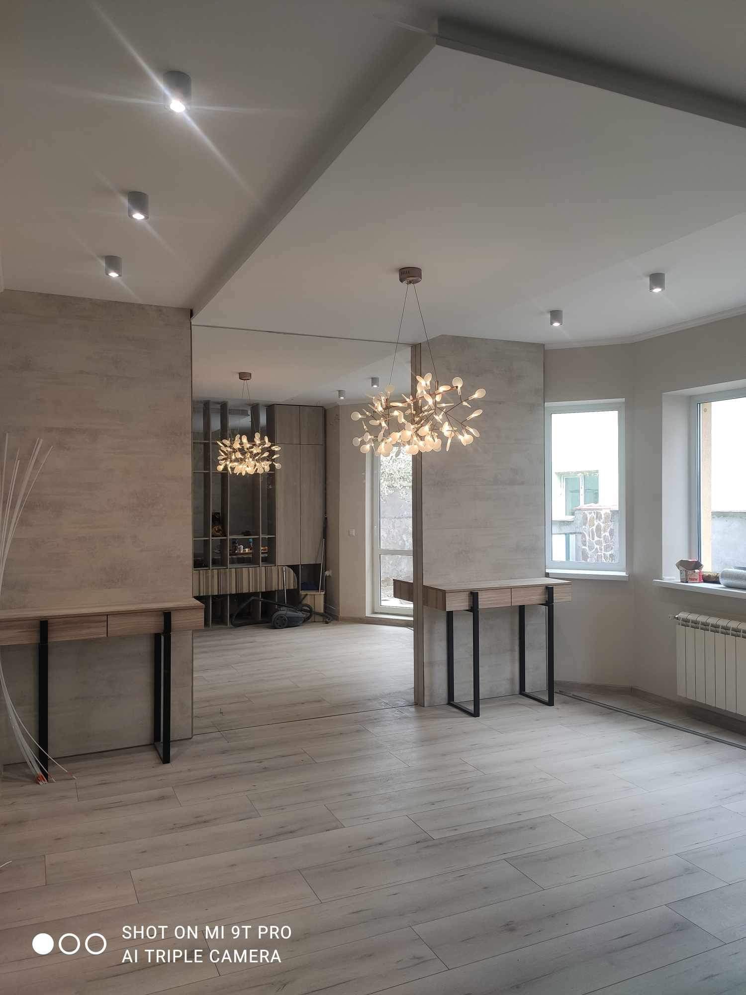 Jasne, przestronne wnętrze po remoncie z drewnianą podłogą, nowoczesnym oświetleniem, konsolami z czarnymi nogami i dużym lustrem na ścianie.