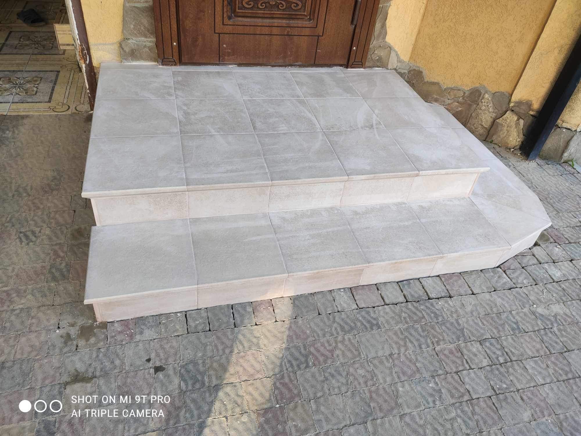 Wyłożone jasnymi płytkami ceramicznymi schody wejściowe do domu, widoczne drzwi wejściowe i fragment elewacji z kamienną okładziną.