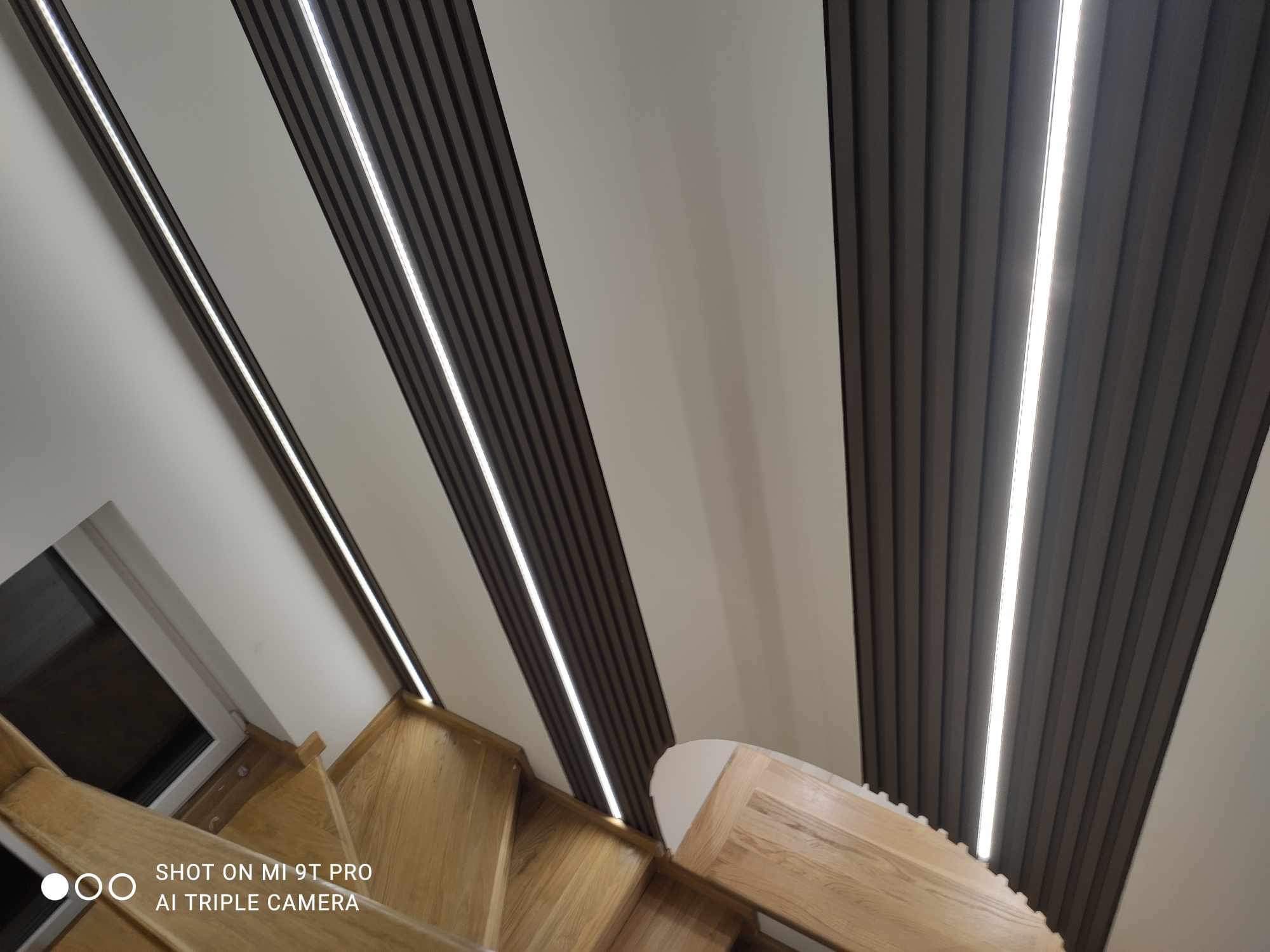 Nowoczesne oświetlenie LED zamontowane wzdłuż drewnianych schodów i dekoracyjnych paneli ściennych w odcieniach bieli i antracytu, widok z dołu.