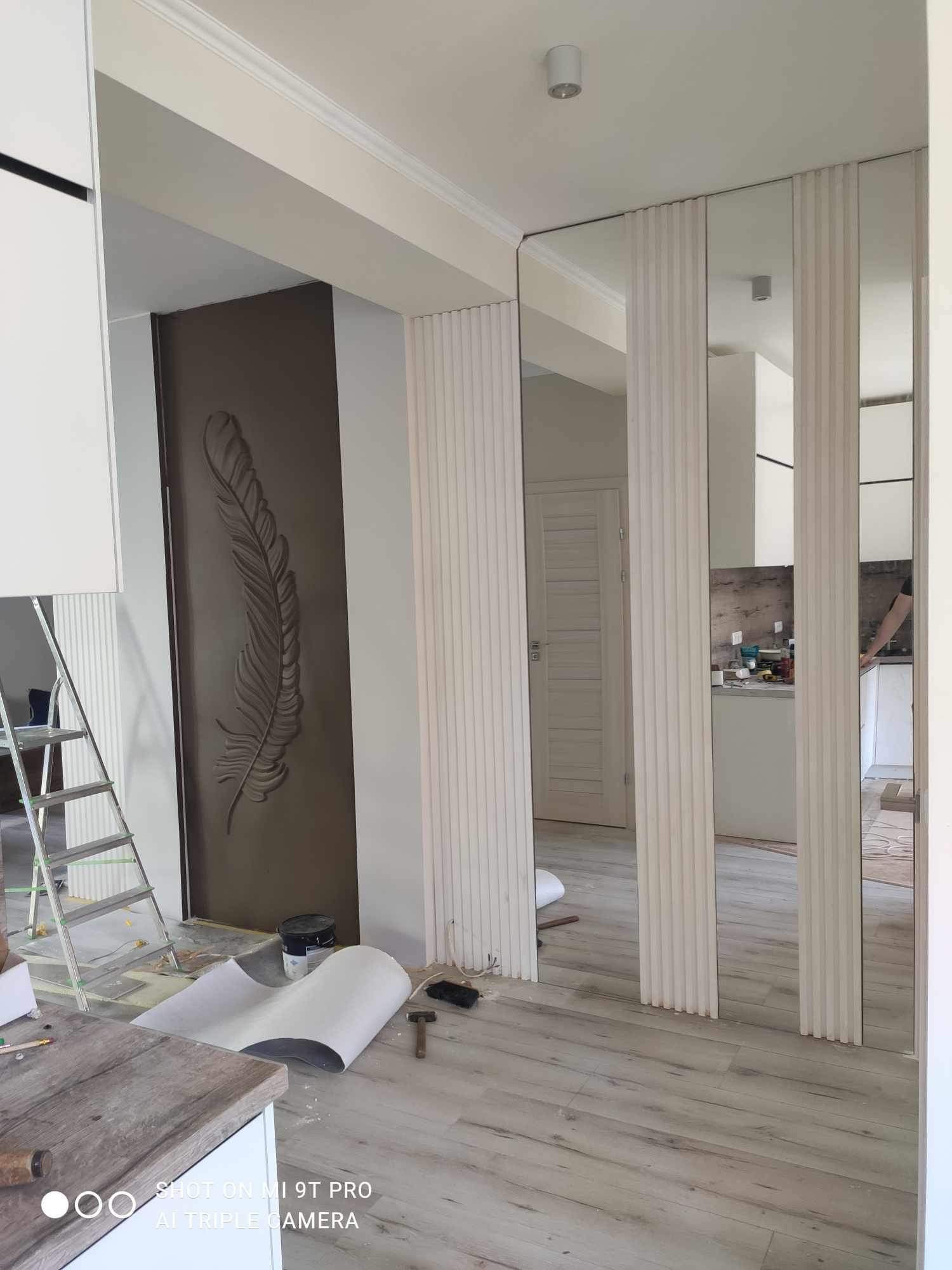 Wnętrze w trakcie wykańczania: widoczna ściana z dekoracyjnym piórem, lustra panelowe, drabina malarska, podłoga i narzędzia budowlane.