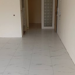 USŁUGI BUDOWLANE RAFAŁ ZUDER - Świeżo położona, błyszcząca, biała podłoga z marmurowym wzorem w pustym pomieszczeniu, widoczne drzwi wejściowe z panelem z luksferów i surowy otwór drzwiowy bez ościeżnicy.