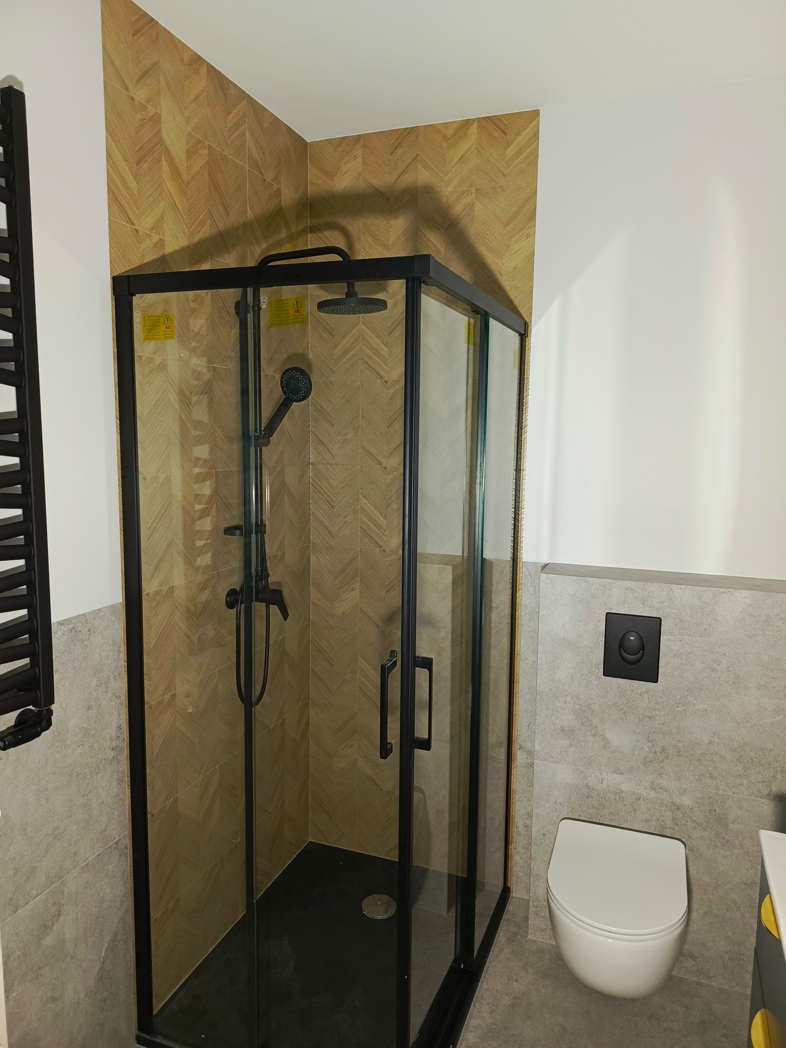 Nowoczesna kabina prysznicowa z czarną ramą, wykończona płytkami imitującymi drewno ułożonymi w jodełkę, w łazience z szarą glazurą i podwieszaną toaletą.