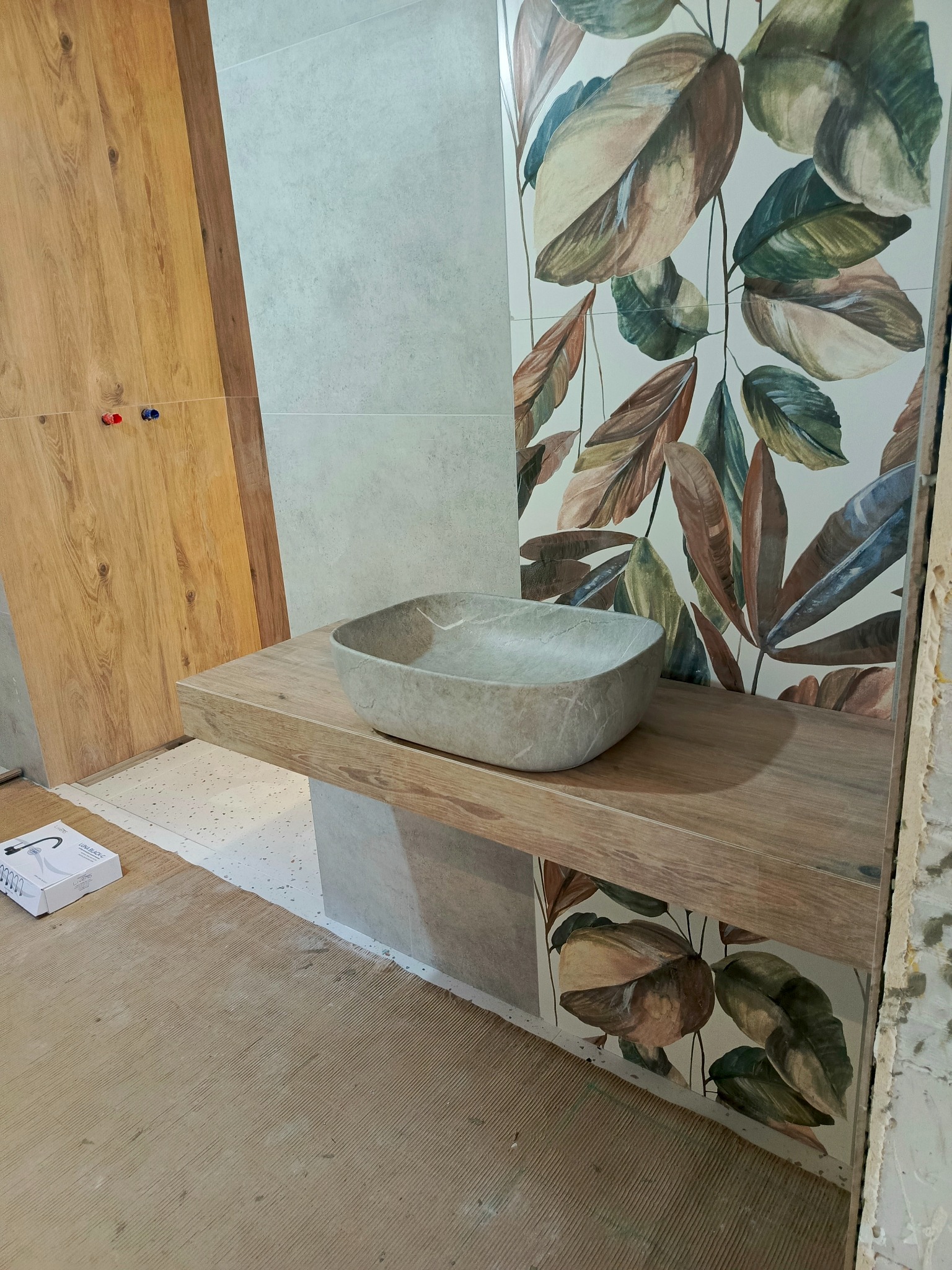 Montaż umywalki nablatowej o nieregularnym kształcie, imitującej kamień, na drewnianym blacie w łazience z płytkami w motywy roślinne. Widoczny karton z armaturą obok blatu.