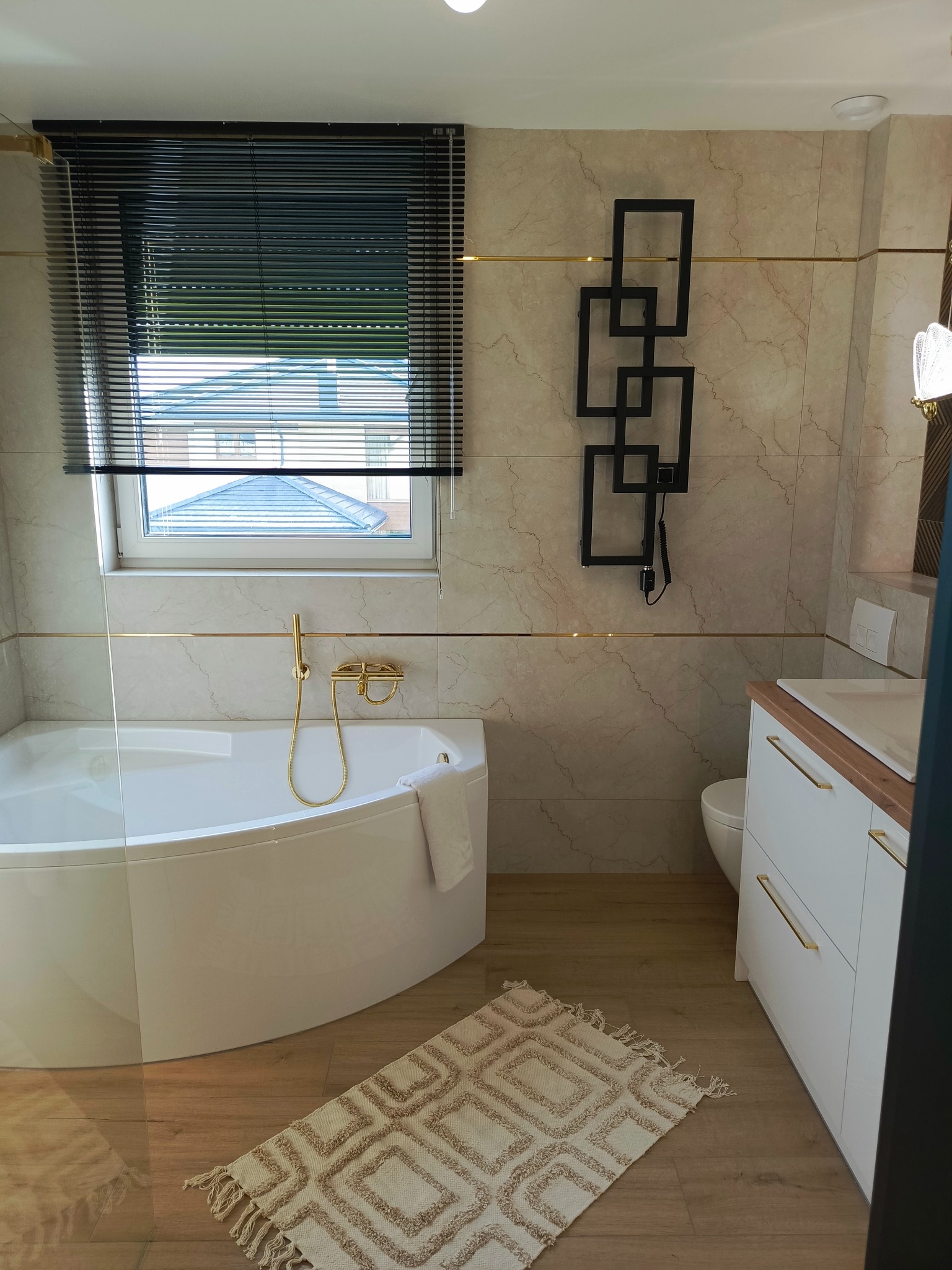 Nowoczesna łazienka z białą wanną narożną, złotą armaturą i beżowymi płytkami imitującymi marmur, uzupełniona czarnym grzejnikiem drabinkowym i dywanikiem z frędzlami.