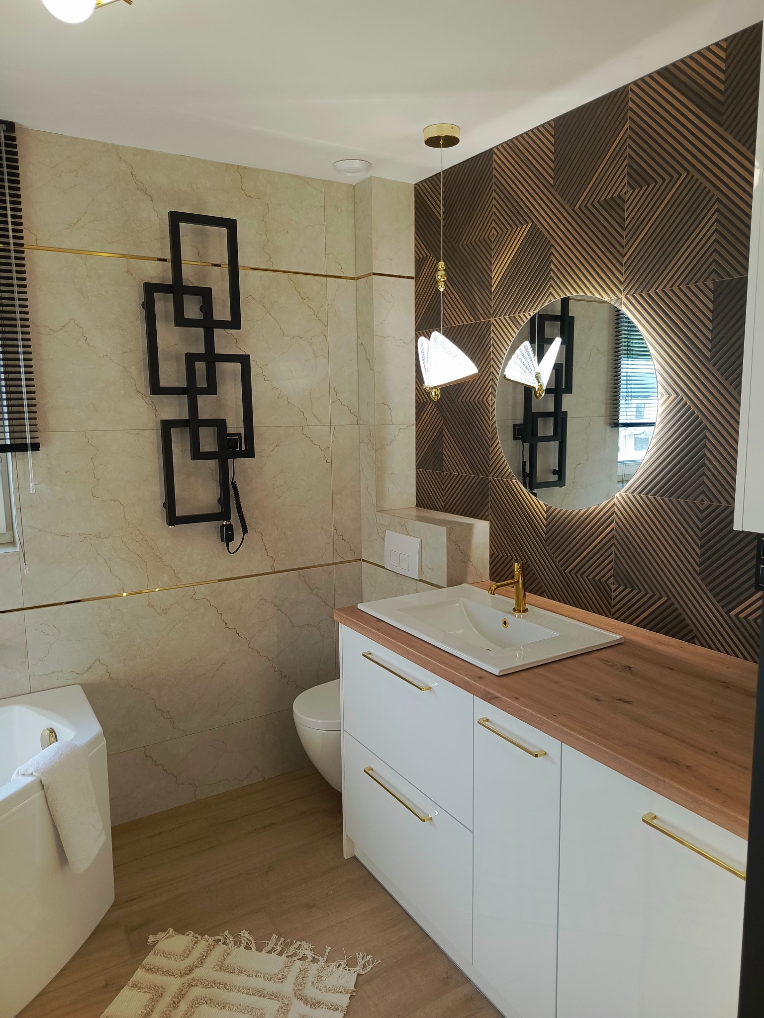 Nowoczesna łazienka z beżowymi marmurkowymi płytkami, geometrycznym grzejnikiem, drewnianą szafką pod umywalką z białymi frontami i złotymi uchwytami oraz dekoracyjną ścianą z drewnianymi panelami...