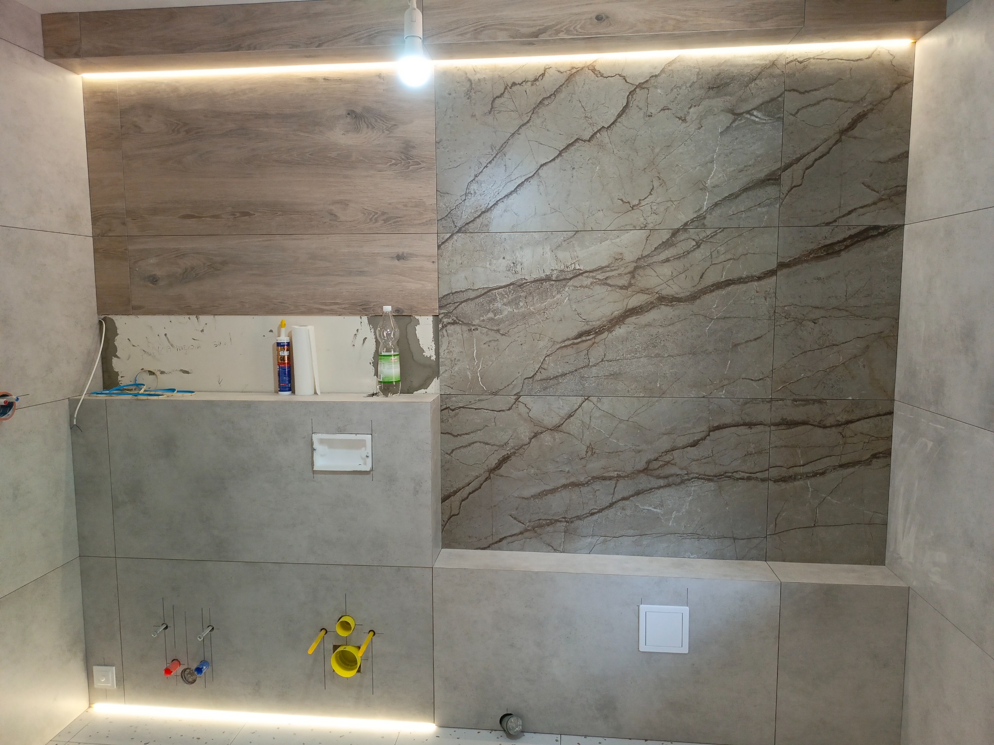 Fragment łazienki w trakcie wykańczania z widocznymi płytkami imitującymi drewno i marmur, podświetleniem LED, instalacją hydrauliczną i niedokończonymi pracami na ścianie.