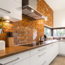 Nowoczesna kuchnia z białymi szafkami, drewnianym blatem i ścianą z czerwonej cegły, oświetlona lampami podszafkowymi i naturalnym światłem z okna.