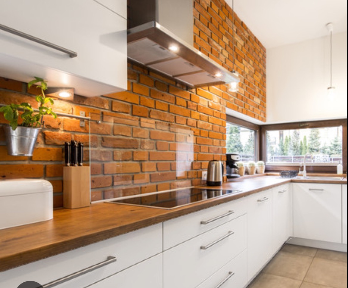 Nowoczesna kuchnia z białymi szafkami, drewnianym blatem i ścianą z czerwonej cegły, oświetlona lampami podszafkowymi i naturalnym światłem z okna.