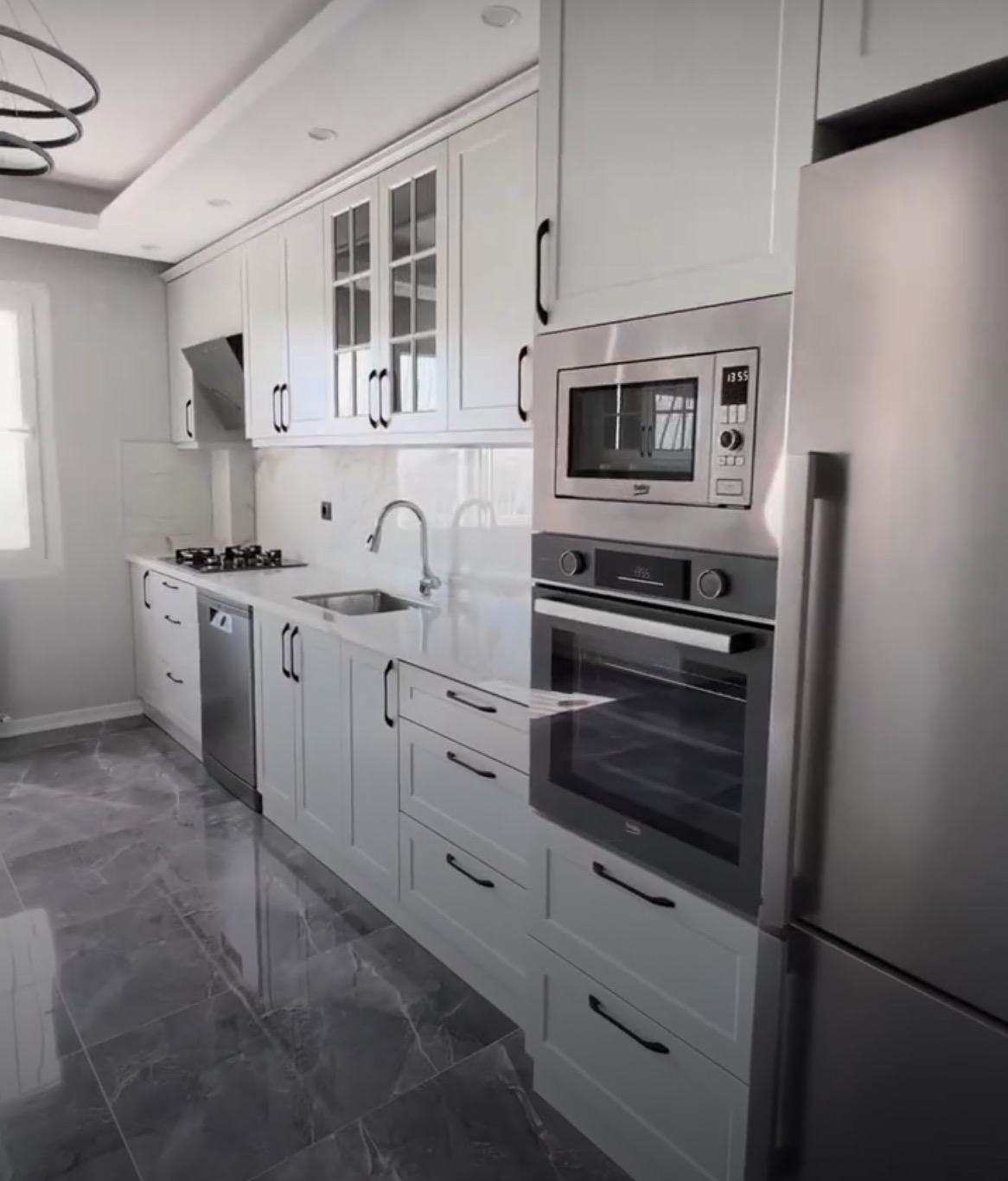 Nowoczesna kuchnia z białymi szafkami z czarnymi uchwytami, marmurowym blatem i szarą marmurową podłogą, z wbudowanym piekarnikiem i mikrofalówką marki Beko oraz stalową lodówką.