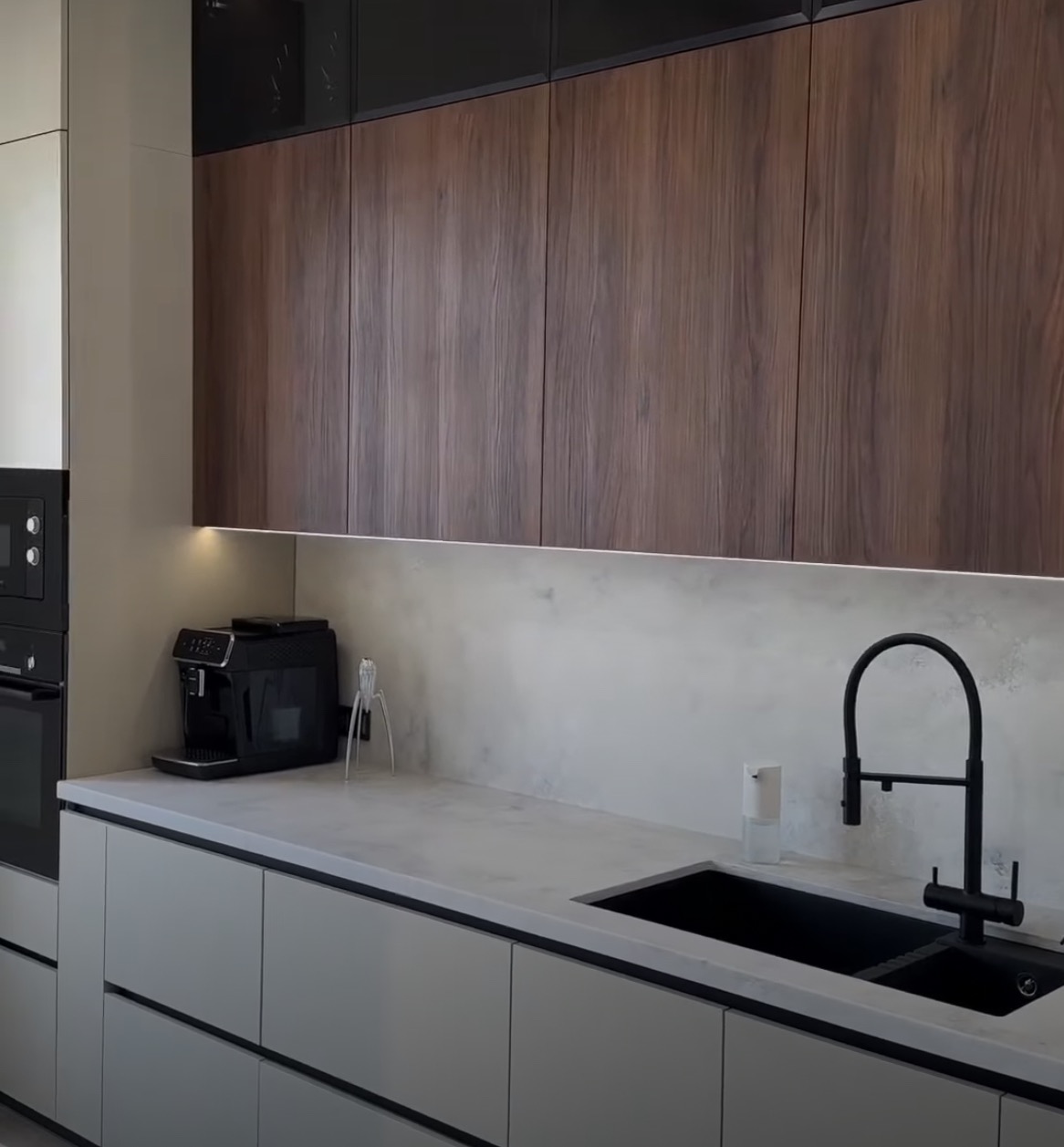 Nowoczesna kuchnia z drewnianymi szafkami górnymi, szarymi szafkami dolnymi, czarnym zlewem i baterią, oraz ekspresem do kawy na blacie.