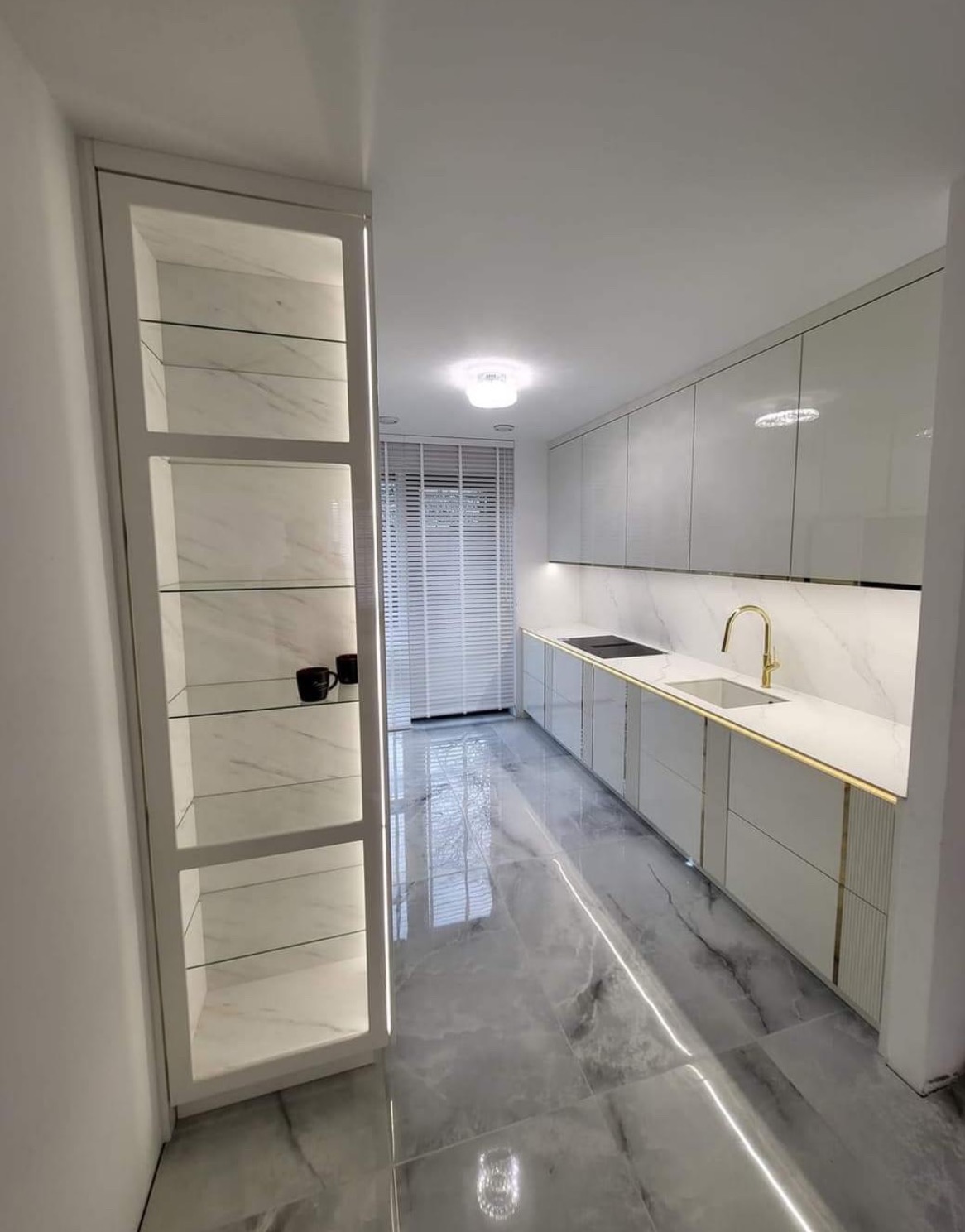 Nowoczesna kuchnia w bieli z marmurowym wykończeniem, złotymi akcentami i przeszkloną witryną z podświetleniem LED, widok na okno z żaluzjami.