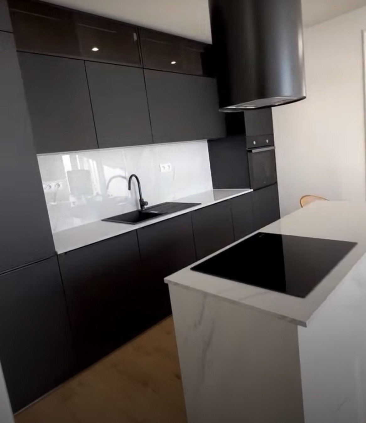 Minimalistyczna kuchnia w czerni i bieli z wyspą kuchenną z blatem imitującym marmur, czarny zlew i bateria, czarna płyta indukcyjna, czarny okap cylindryczny.