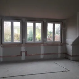 Surowe wnętrze poddasza z trzema oknami i instalacją miedzianą na ścianach przed wykończeniem, widoczne łączenia płyt gipsowo-kartonowych.