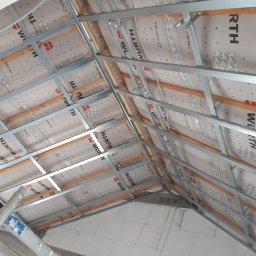 CNG-Uslugi Budowlane - Konstrukcja poddasza z metalowym stelażem i membraną izolacyjną Wurth, widok z dołu, częściowo ocieplone ściany z białych bloczków.