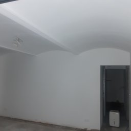 CNG-Uslugi Budowlane - Wnętrze pokoju po malowaniu ścian na biało, z widocznym sufitem z zaokrąglonymi krawędziami, wystającymi przewodami elektrycznymi i osuszaczem powietrza w przejściu do innego pomieszczenia.
