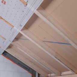 CNG-Uslugi Budowlane - Konstrukcja drewniana poddasza z widoczną membraną dachową z logo Würth, zamocowana do belek stropowych, w tle ściana z białych bloczków.