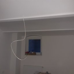 CNG-Uslugi Budowlane - Wnętrze w trakcie prac wykończeniowych: pomalowane ściany na biało, widoczne listwy przysufitowe, okno z zabezpieczeniem, zwisający kabel oświetleniowy i grzejnik pod oknem.