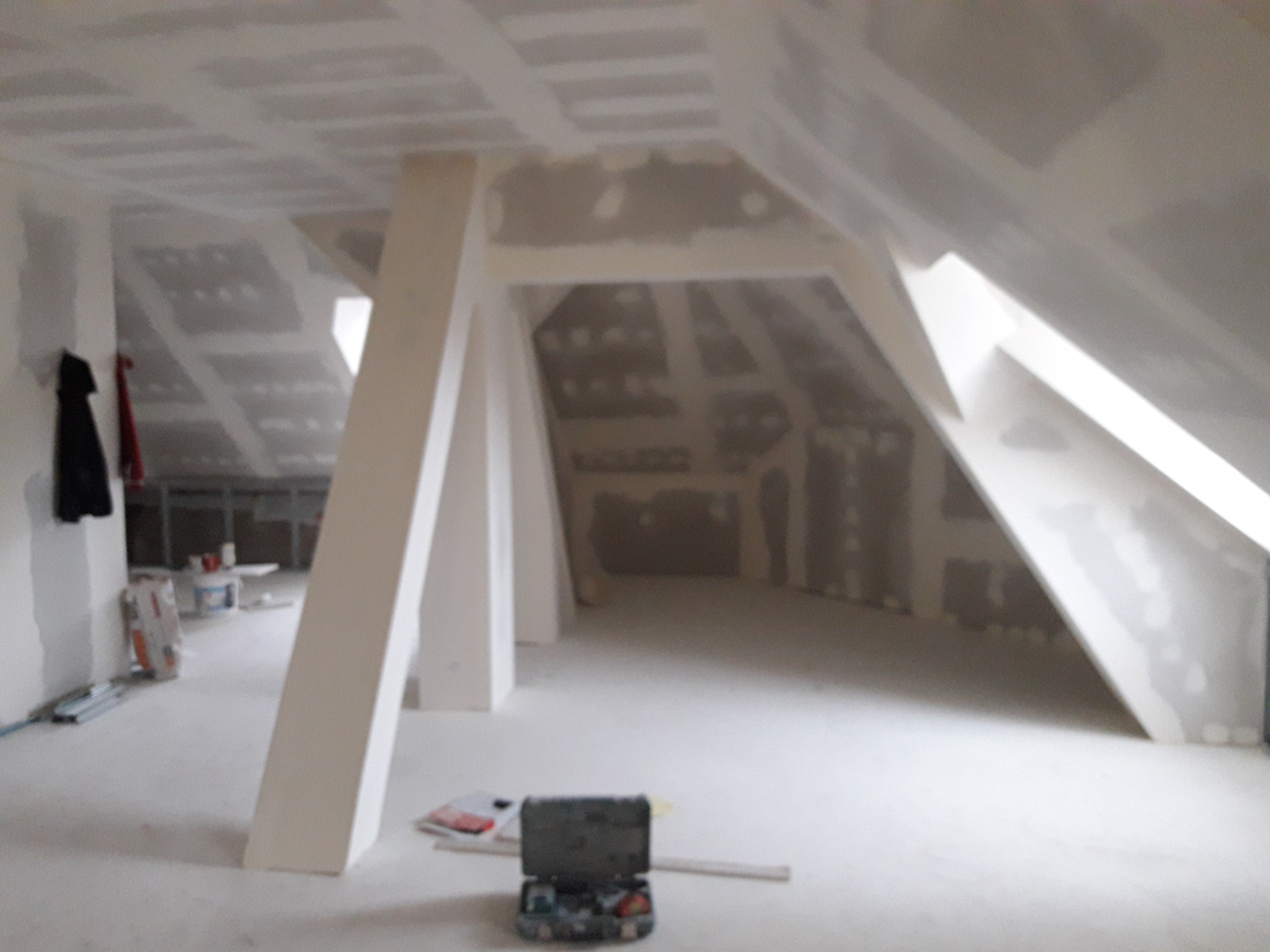 Poddasze w trakcie wykańczania: widoczne płyty gipsowo-kartonowe na ścianach i suficie, szpachlowane łączenia, białe słupy konstrukcyjne, skrzynka z narzędziami na podłodze.