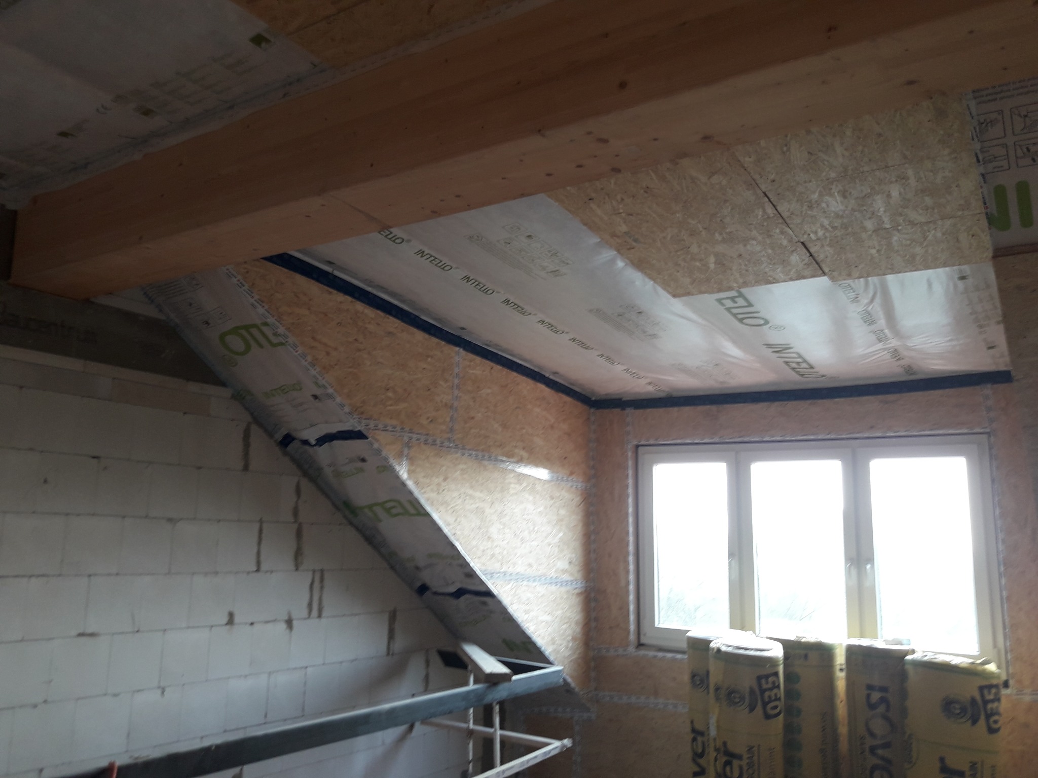 Wnętrze poddasza w trakcie ocieplania: widoczne belki drewniane, płyty OSB, folia paroizolacyjna INTELLO, bloczki z betonu komórkowego oraz paczki wełny mineralnej ISOVER przy oknie.