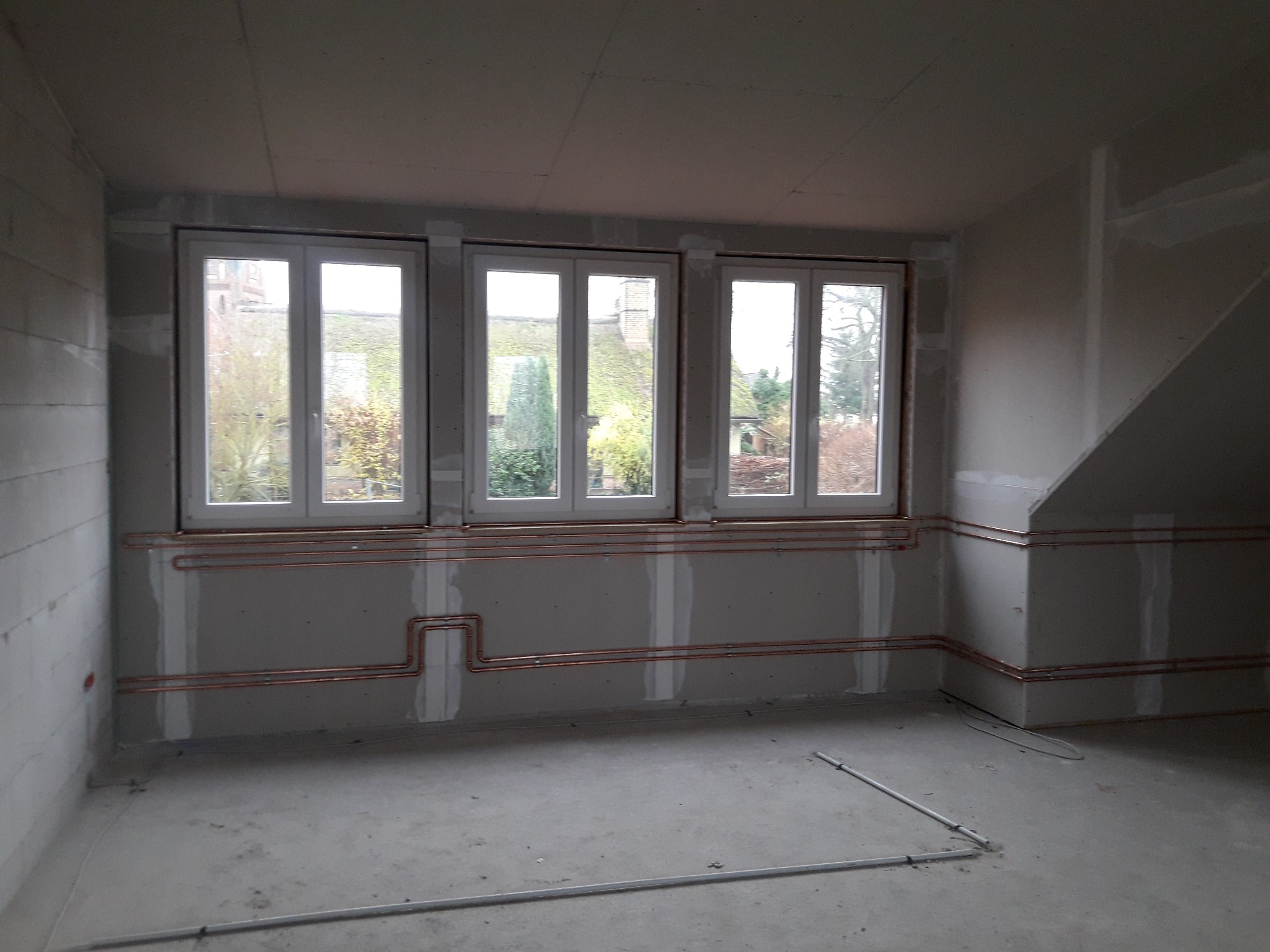 Surowe wnętrze poddasza z trzema oknami i instalacją miedzianą na ścianach przed wykończeniem, widoczne łączenia płyt gipsowo-kartonowych.