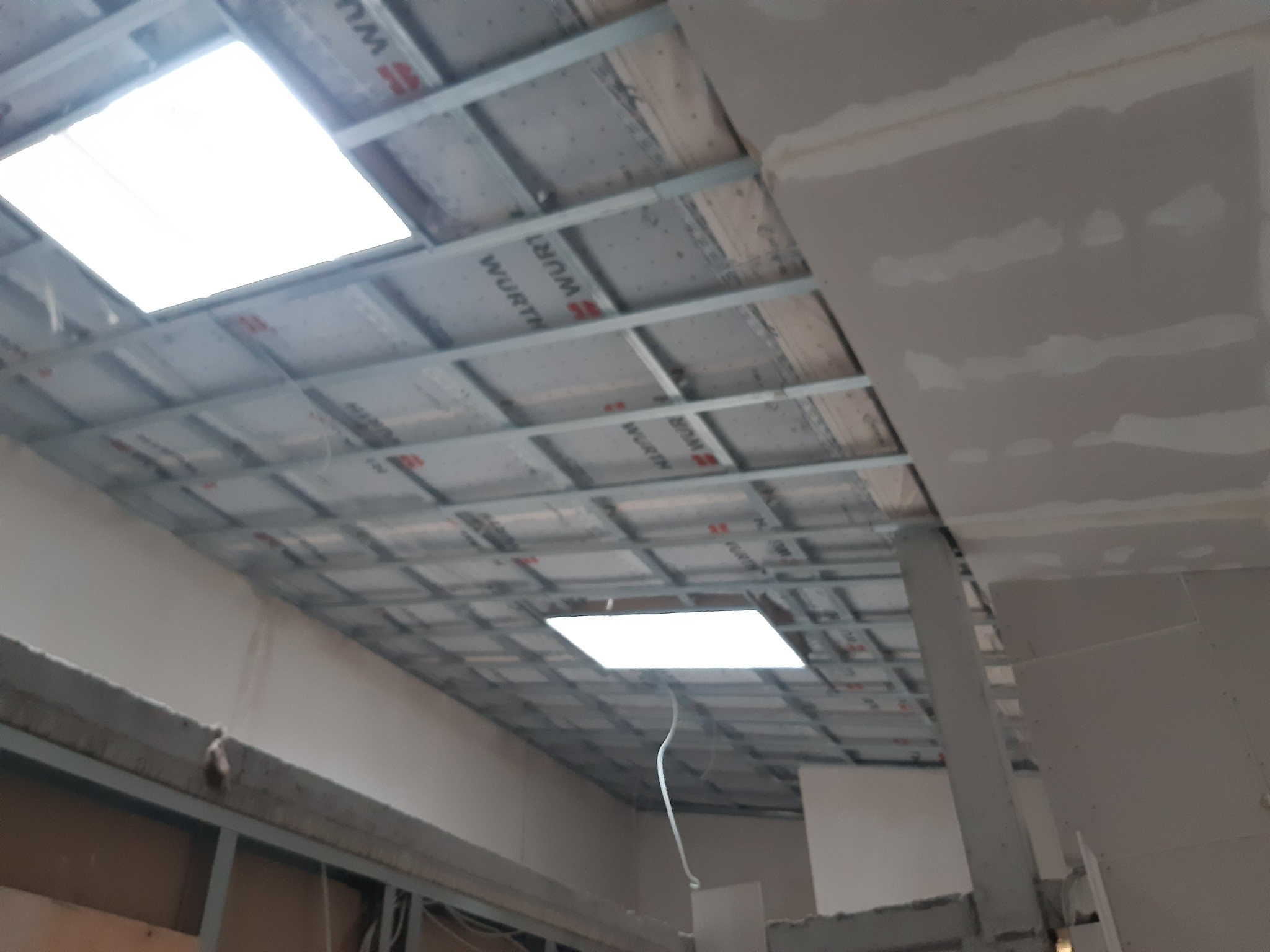 Sufit w trakcie montażu z widoczną konstrukcją metalową, izolacją z nadrukiem Wurth oraz fragmentami płyt gipsowo-kartonowych, oświetlony naturalnym światłem z okien dachowych.