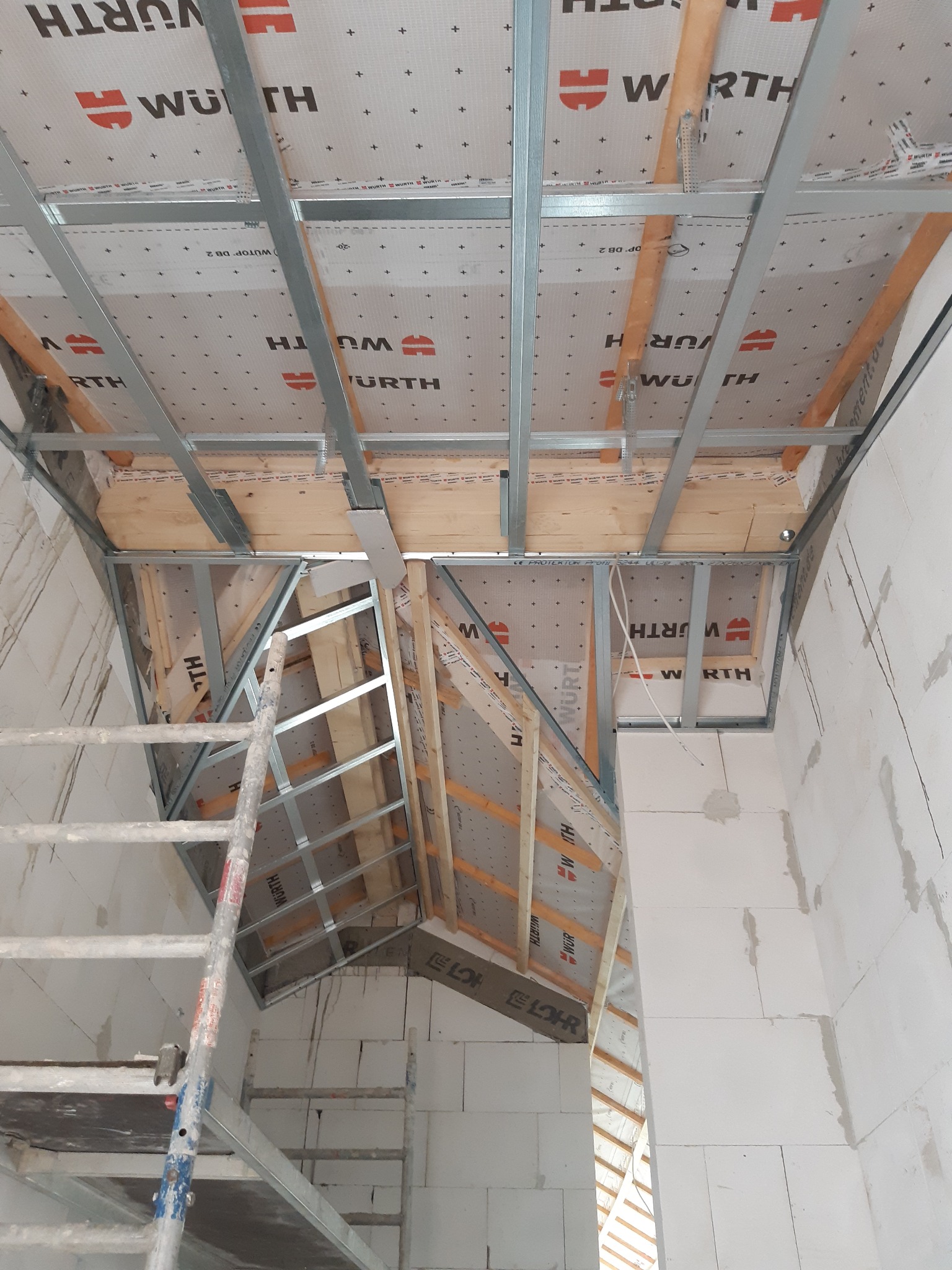 Szkieletowa konstrukcja poddasza z widocznymi elementami drewnianymi i metalowymi profilami, pokryta membraną marki Wurth z drabiny podczas montażu.