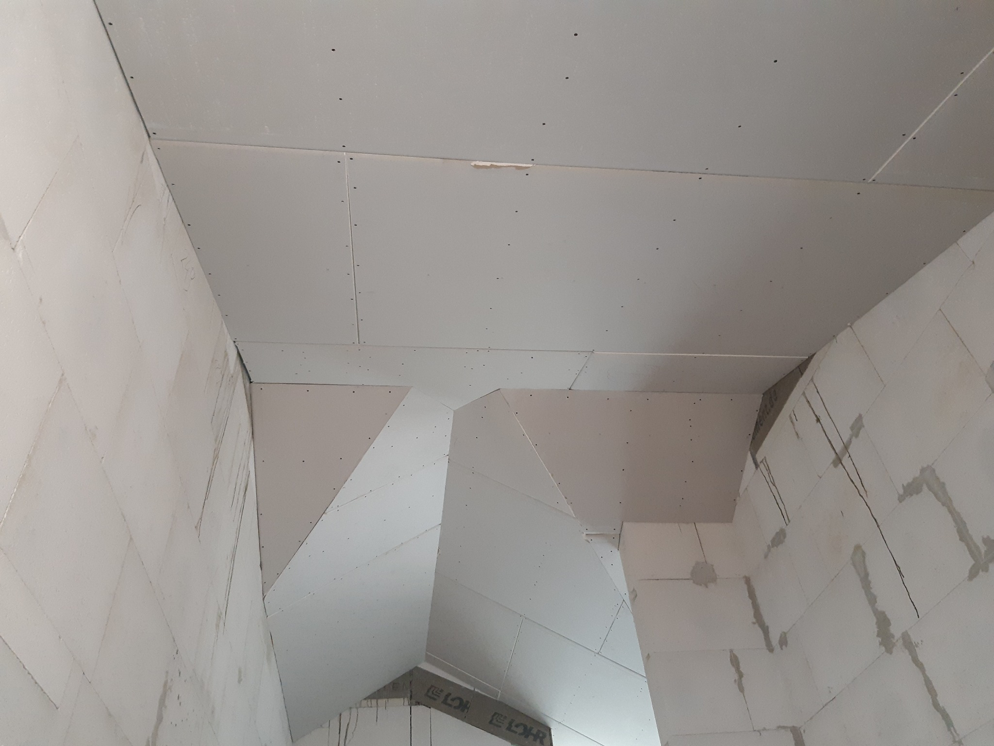 Sufit i ściany pokryte płytami gipsowo-kartonowymi w surowym stanie, widoczne ślady łączeń i szpachlowania, blok konstrukcyjny na ścianie, widok z dołu.
